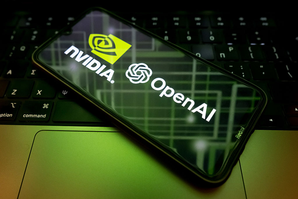 The Nvidia and OpenAI logos are seen on a phone screen in front of a laptop with green lighting.