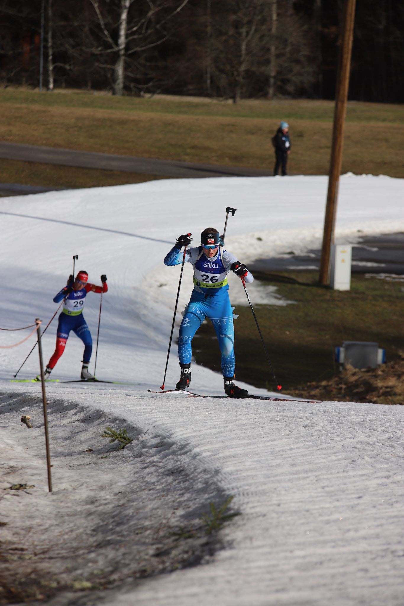 Two biathletes race on a narrow strip of snow with grass and mud on either side.
