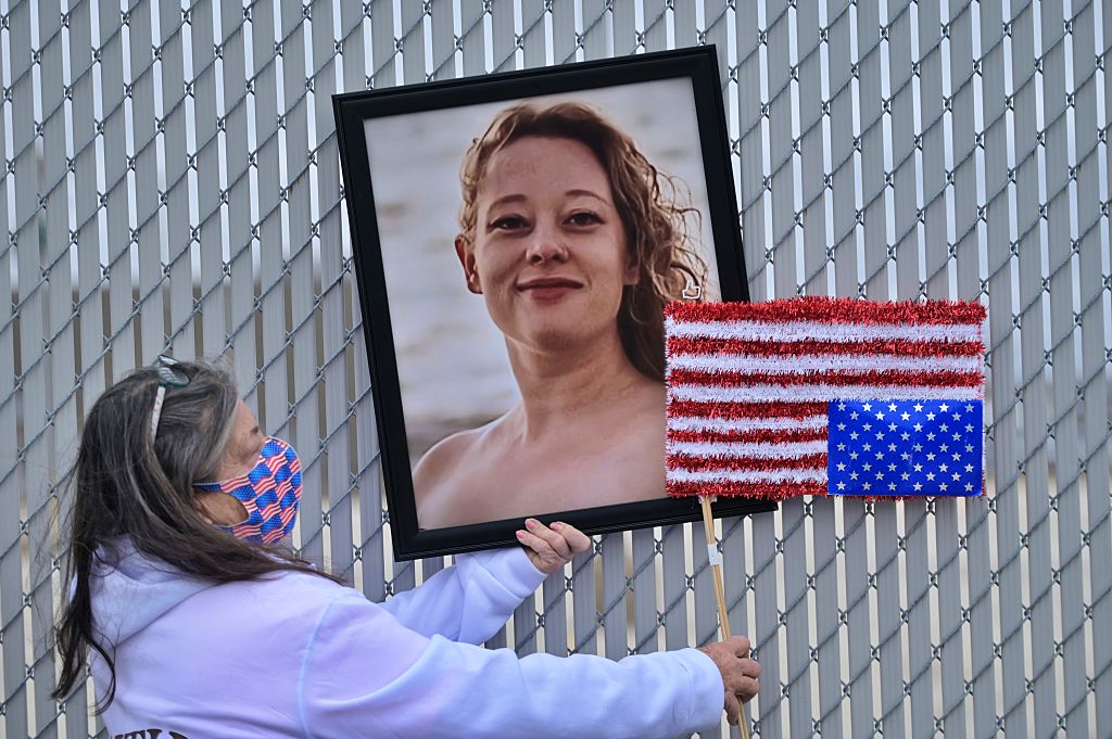 A woman in a mask holds up a portrait of Renee Nicole Good and an upside down American flag in front of a chain link fence.