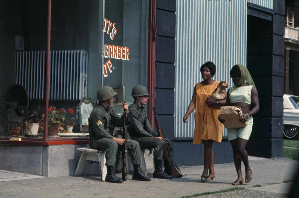 Two women walk past National Guardsmen seated on a bench outside a shop during rioting in Detroit, Mich., July 27 1967.